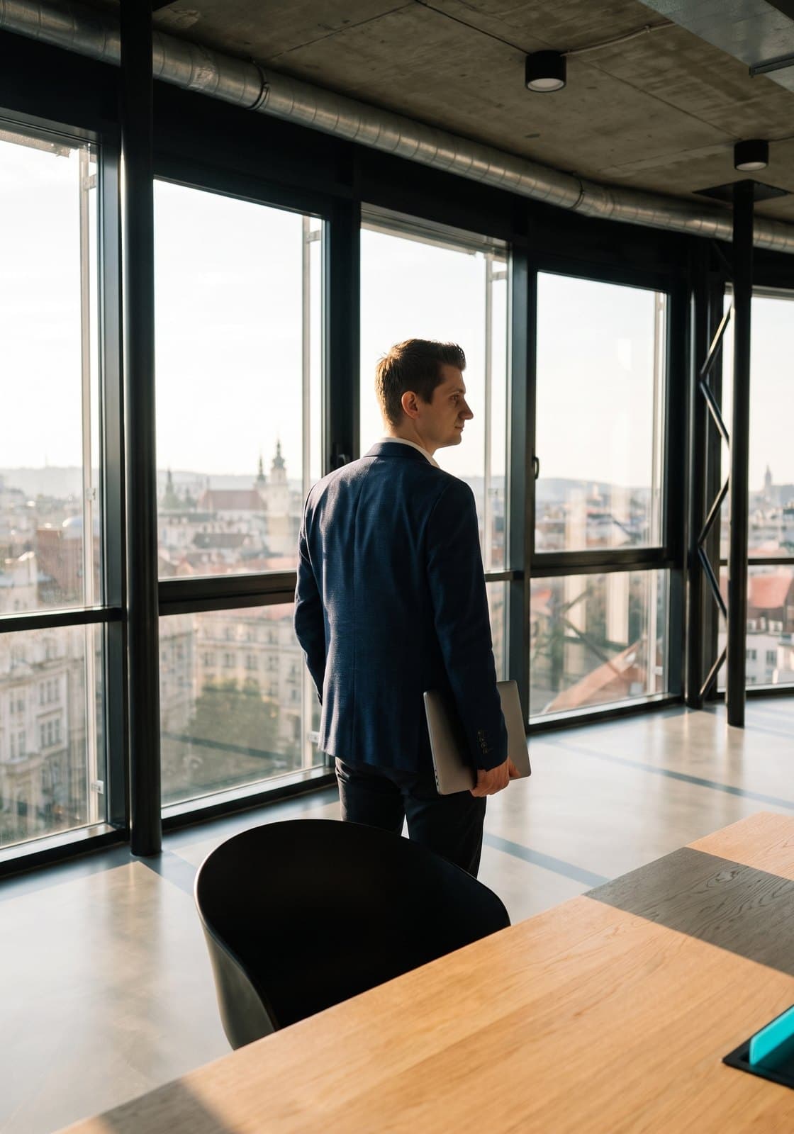 A man in a navy suit holding a laptop, looking out over the city from a corner office window